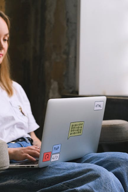 A person wearing a white T-shirt and blue jeans is kneeling on the floor inside a property, using a red marker to write 'Spares' on the top of a cardboard box that is sealed with white packing tape. S