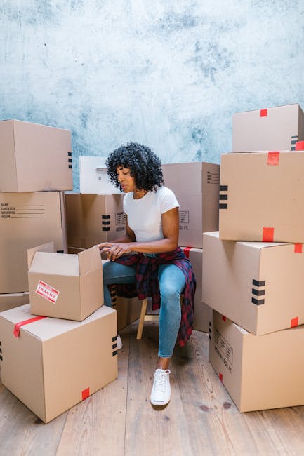 A woman with curly black hair, wearing a white T-shirt, blue jeans, and white sneakers, is seated on a wooden chair among numerous cardboard boxes of various sizes, some with black tape strips and red