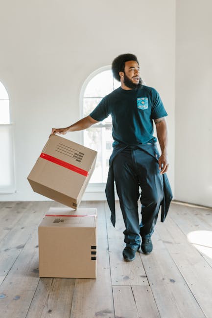 A woman with curly black hair, wearing a white T-shirt, blue jeans, and white sneakers, is seated on a wooden chair among numerous cardboard boxes of various sizes, some with black tape strips and red